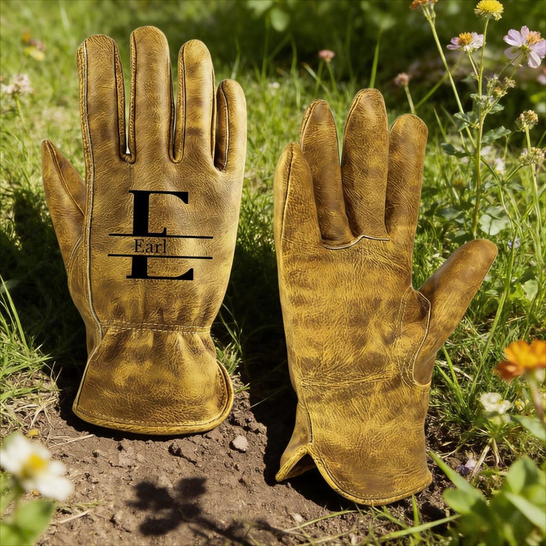 May include: A pair of brown leather gardening gloves with the name "Earl" and the letter "E" printed in black. The gloves are sitting on the ground in a garden setting, surrounded by grass and flowers.