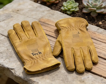 Puede incluir: Un par de guantes de trabajo de cuero color canela con la palabra "Tech" impresa en el guante izquierdo. Los guantes están sobre una superficie de madera junto a una superficie de piedra con suculentas en el fondo. El guante derecho tiene la letra "L".