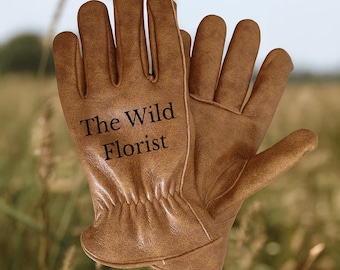 May include: A pair of brown leather gardening gloves with the text "The Wild Florist" printed on the front. The gloves are designed for outdoor work and feature a classic design with reinforced stitching.