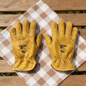 Puede incluir: Un par de guantes de trabajo de cuero amarillo con un logotipo personalizado con el nombre "Christina". Los guantes están colocados sobre un paño a cuadros marrón y blanco, lo que sugiere jardinería o actividades al aire libre.