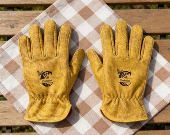 Puede incluir: Un par de guantes de trabajo de cuero amarillo con un logotipo personalizado con el nombre "Christina". Los guantes están colocados sobre un paño a cuadros marrón y blanco, lo que sugiere jardinería o actividades al aire libre.