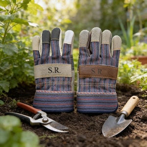 May include: A pair of gardening gloves with blue and red striped fabric, tan leather accents, and the initials "S.R." on the wrist straps. A small trowel and pruning shears are in the foreground. The gloves are designed for gardening.