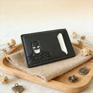 Pode incluir: Porta-cartões de couro preto com design do Batman. O porta-cartões tem interior branco e costuras brancas. É exibido em uma bandeja de madeira com um pano de serapilheira.
