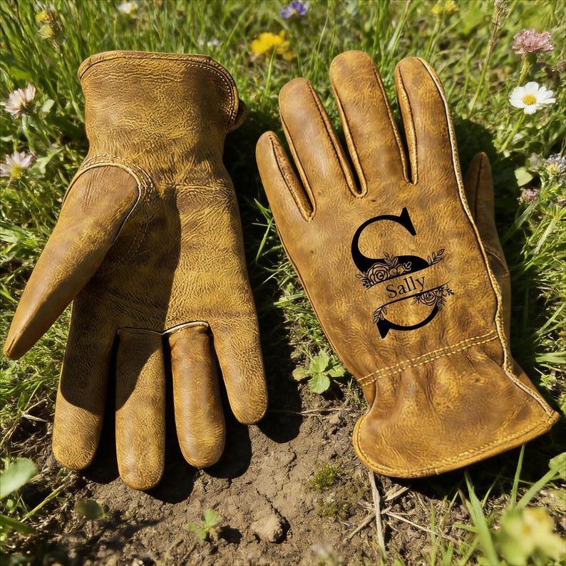 May include: A pair of brown leather work gloves. One glove features the initial "S" with the name "Sally" in a floral design. The gloves are lying on the ground in a grassy area.
