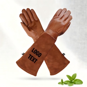 May include: Brown leather gardening gloves with long sleeves, featuring the text "LOGO TEXT" in black. The gloves are designed for protection and have a stitched seam detail. A small sprig of mint is in the bottom right corner.