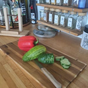 Può includere: Un tagliere rettangolare in legno con sopra un peperone rosso intero, un cetriolo intero, un cetriolo parzialmente affettato e un coltello da cucina con manico in legno. Il tagliere è posizionato su un piano di lavoro in legno.