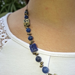 Può includere: Primo piano di una collana con perline quadrate e rotonde blu e macchiate. La collana ha un cordino scuro e piccoli distanziatori color ottone. La collana è indossata su una camicia bianca.