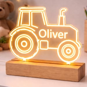 Puede incluir: Un letrero de luz de neón LED con forma de tractor y un brillo amarillo cálido, que presenta el nombre Oliver en letras blancas. El contorno del tractor de acrílico está montado sobre una base rectangular de madera clara con un cable.