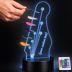 May include: A guitar headstock-shaped LED lamp with blue glowing outlines, featuring the text "your custom text" and six colourful guitar picks arranged in a vertical line. The lamp sits on a black circular base and includes a remote control.