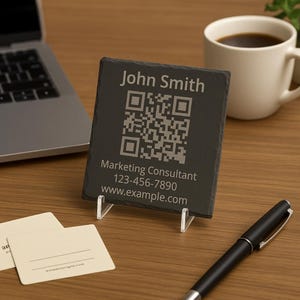 Op de afbeelding: Een zwarte leistenen visitekaarthouder met een QR-code en de naam "John Smith" in het wit gegraveerd. De tekst "Marketing Consultant", een telefoonnummer en een websiteadres worden ook weergegeven. De kaart wordt ondersteund door een transparante plastic standaard.