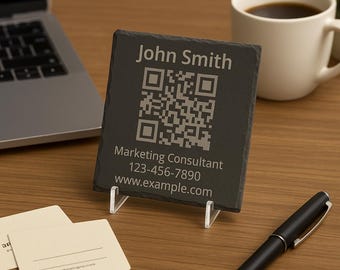 Op de afbeelding: Een zwarte leistenen visitekaarthouder met een QR-code en de naam "John Smith" in het wit gegraveerd. De tekst "Marketing Consultant", een telefoonnummer en een websiteadres worden ook weergegeven. De kaart wordt ondersteund door een transparante plastic standaard.