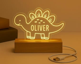 Könnte beinhalten: Ein leuchtendes Nachtlicht in Dinosaurierform mit dem Namen "OLIVER" auf dem Körper. Das Licht besteht aus klarem Acryl und steht auf einem Holzsockel. Ein USB-Kabel ist mit dem Sockel verbunden.