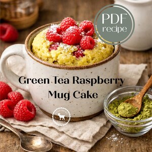 May include: A mug cake with green tea and raspberries. The cake is in a speckled white mug, topped with fresh red raspberries and powdered sugar. A small bowl of green matcha powder and a wooden spoon are next to the mug. Text on the mug says "Green Tea Raspberry Mug Cake."