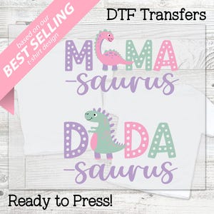 以下が含まれることがあります： 「MAMA-saurus」と「DADA-saurus」の文字と、ピンク、紫、緑色の恐竜グラフィックがプリントされたDTF転写の白いTシャツ。画像には「Best Selling」と「Ready to Press!」の文字も含まれています。