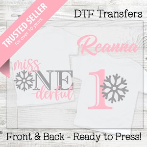 May include: White t-shirts with DTF transfers. One shirt features the text "miss ONE-derful" with a snowflake graphic. The other shirt has the name "Reanna" and the number "1" with a snowflake. The text "Front & Back - Ready to Press!" is also visible.