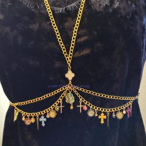 May include: A gold-toned body chain featuring a central clover-shaped connector, multiple draped chains, and various dangling charms including small crosses, floral motifs, and pointed spikes. The piece is displayed against a dark velvet fabric.