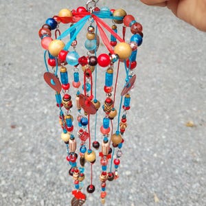 May include: A hanging mobile ornament constructed from a circular wire frame with multiple dangling strands of colorful beads. The beads are made of wood, glass, and plastic in shades of red, blue, and tan, interspersed with thin ribbons and metal rings.