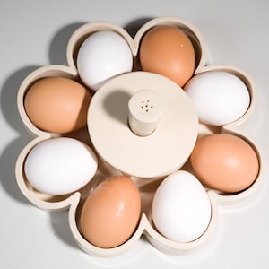 May include: A white egg holder shaped like a flower with six compartments. The holder is filled with six eggs, three white and three brown.