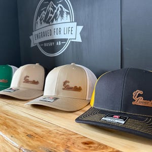 May include: Four baseball caps on display, each with a different colour combination. The caps have a curved peak and a mesh back. The first cap is green, the second and third are tan, and the fourth is black with a yellow mesh back. Each cap has a leather patch that says "Loose Ends".