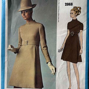 May include: A Vogue Americana sewing pattern envelope 2068 featuring Teal Traina designs. The cover displays a tan long-sleeved dress with a high collar and matching belt, alongside a brown short-sleeved dress with a buckled belt illustration.