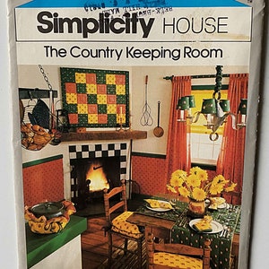 May include: A Simplicity House pattern for a country-style room. The pattern shows a fireplace with a checkered tile surround, a wooden table with chairs, and a quilt hanging on the wall. The text on the pattern reads "The Country Keeping Room".