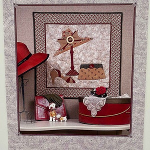 May include: A quilt pattern titled "Rendezvous in the Park" by Elle Colquitt. The pattern features a red hat, a red and white hatbox, a red purse, and two white dogs. The quilt is framed by a white picket fence.