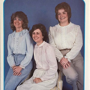 May include: Three women wearing white and light blue blouses with lace trim. The women are sitting on a chair with a striped pattern. The image is from a sewing pattern booklet for a machine-sewn French blouse. The text on the image reads "#13 Brenda Olivia Machine French Sewn Blouse".