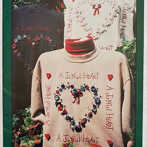 May include: A white sweatshirt with a red heart design made of buttons. The heart is surrounded by the text "A Joyful Heart". The sweatshirt is on a mannequin and the background is a green bush.