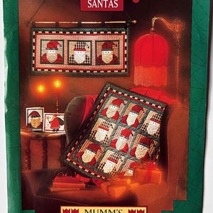 May include: A Christmas-themed image featuring a quilt and wall hanging with Santa Claus designs. The quilt is square, with multiple Santa faces. The wall hanging has three Santa faces. The image also includes a red lamp and gifts.