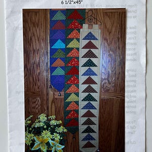 May include: A colorful, handmade bellpull with a "Flying Geese" design. The bellpull is approximately 6.5 inches wide and 45 inches long. It features rows of triangular fabric pieces in various colors, including blue, red, green, and yellow. The bellpull hangs from a wooden door.