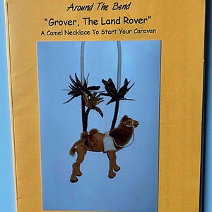 May include: A brown camel with a white saddle and a palm tree necklace. The camel is a cloth animal design by Ruth Prest. The text "Grover, The Land Rover" and "A Camel Necklace To Start Your Caravan" are printed on the front of the booklet. The text "cloth animal design by Ruth Prest © 1999/ AB103" is printed on the bottom of the booklet.