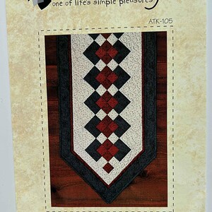 May include: A Seminole table runner pattern with a geometric design in red, black, and white. The pattern features diamond shapes and is framed by a dark border. The text "Atkinson Designs" and "SEMINOLE TABLE RUNNER" are visible.