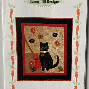 May include: A quilting pattern featuring a black cat with a red heart and a broom, surrounded by red and green leaves. The pattern is titled "Postcard Kitty... 1027 A Topsy Turvy Pattern".