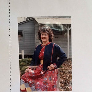 May include: A woman wearing a blue denim jacket with red trim and a red floral dress with a yellow belt. The jacket has hidden pockets. The photo was taken in front of a wooden building. The text "DENIM JACKET with HIDDEN POCKETS" is at the top of the image. The text "--- by: JEANNE VEHR HAMBLETON 1986" is at the bottom of the image.