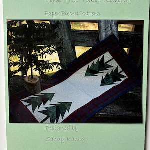 May include: A paper pieced pattern for a pine tree table runner. The runner features a white center with green pine tree shapes, framed by a red and blue plaid border. The text "Pine Tree Table Runner" and "Designed by Sandy Kalvig" are visible.