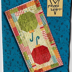 May include: A quilted table runner with umbrella designs in green and red, featuring a patchwork design with various patterns and colors. The text "pieces from my heart" is visible. The pattern is titled "#350 TABLE ATTIRE".