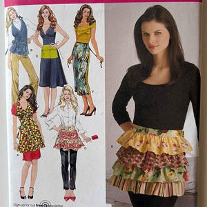 May include: Simplicity sewing pattern 3752 featuring apron designs. The pattern includes various apron styles, with tiered ruffles, floral prints, and a classic design. The image also shows a woman wearing a tiered ruffle apron.