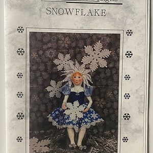 May include: A 16-inch seated doll pattern with button jointed legs and arms. The doll is dressed in a blue dress with white snowflakes and a white snowflake crown. The background is a dark brown with white snowflakes.