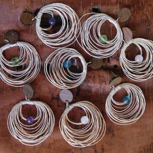 May include: A selection of handmade earrings with a circular design. Each earring has a wooden stud, a wrapped white cord design, and a small, colourful bead. The earrings are displayed on a textured, brown surface.