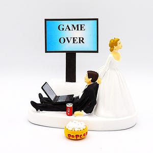 May include: A wedding cake topper featuring a bride pulling her groom away from a laptop. The groom is wearing a black suit and is sitting down with a can of cola and a bowl of popcorn. A sign above the groom reads "GAME OVER".