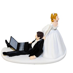 May include: A wedding cake topper featuring a bride in a white gown pulling a groom in a black suit who is engrossed in a laptop. The figures are on a white base, a humorous take on modern relationships.