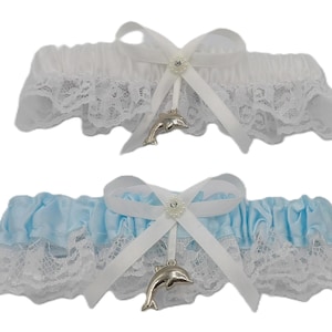 May include: Two garters, one white and one light blue, with white lace trim. Each garter has a white bow with a rhinestone center and a silver dolphin charm. The garters are likely for a wedding or special occasion.