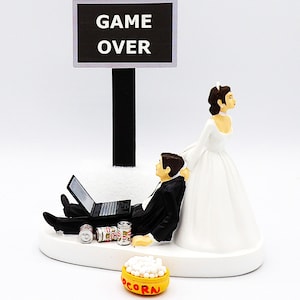 May include: A humorous wedding cake topper featuring a bride and groom. The groom is seated with a laptop and cans, while the bride stands beside him. A sign reads "GAME OVER", and a popcorn bowl is in front.