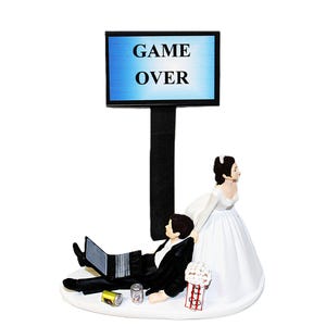 May include: A wedding cake topper featuring a bride and groom. The groom is lounging with a laptop and drinks, while the bride stands beside him. A sign that reads "GAME OVER" is above them. The cake topper is white and black.