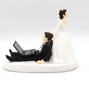 May include: Wedding cake topper featuring a bride in a white gown and a groom in a black suit, reclining with a laptop. The base is white, and the figures are detailed.
