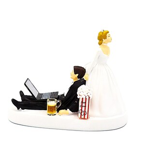 May include: A humorous wedding cake topper featuring a bride and groom. The groom is lounging with a laptop and beer, while the bride looks on. A popcorn container is also included. The figures are on a white base.