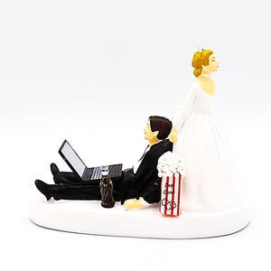 May include: A wedding cake topper featuring a bride in a white gown and a groom in a tuxedo, lounging with a laptop and a bottle. A popcorn container is also present. The theme is a humorous take on modern relationships.