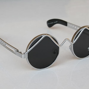 May include: Silver round sunglasses with black lenses and a unique, angular frame design.