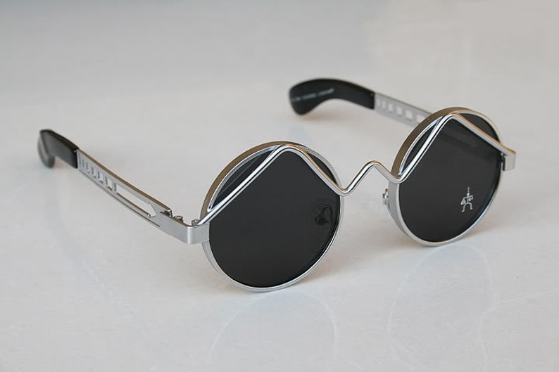 May include: Silver round sunglasses with black lenses and a unique, angular frame design.