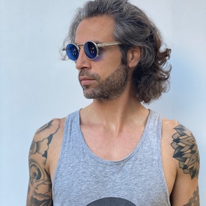 May include: A man with long gray hair wearing round blue sunglasses with a silver frame. He is wearing a gray tank top with a black circle and a geometric design on the front. He has tattoos on both arms.